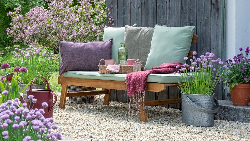 © Gartenbildagentur Friedrich Strauss Ein gemütlicher Gartenplatz mit Holzbank, Kissen und Decke ist von blühenden Pflanzen und Frühlingsblumen umgeben.