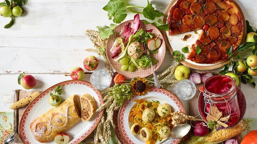 Ein herbstlich gedeckter Tisch zeigt verschiedene regionale Speisen, Äpfel, Kürbisse und eingelegtes Gemüse.