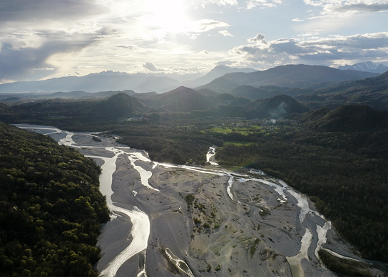Eine weite Landschaft zeigt einen verzweigten Flusslauf im Tagliamento, umgeben von Hügeln und Bergen unter bewölktem Himmel.