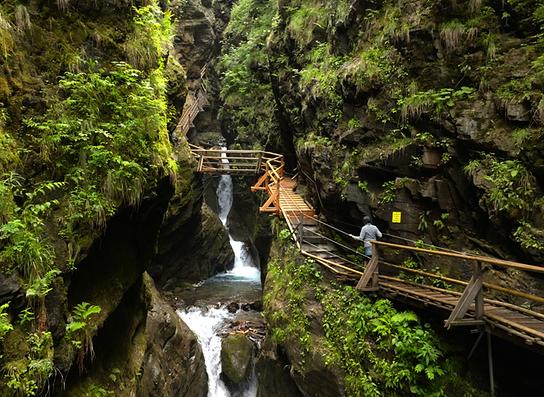 Ein schmaler Steg führt durch eine moosbewachsene Schlucht mit kleinen Wasserfällen.