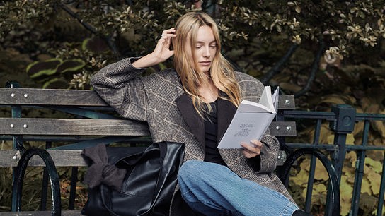 Eine Frau in einem langen Mantel und Jeans sitzt mit einem Buch auf einer Parkbank und liest entspannt.