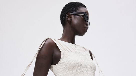 Ein dunkelhäutiges Model trägt ein elegantes, cremeweißes Kleid und eine dunkle Sonnenbrille.