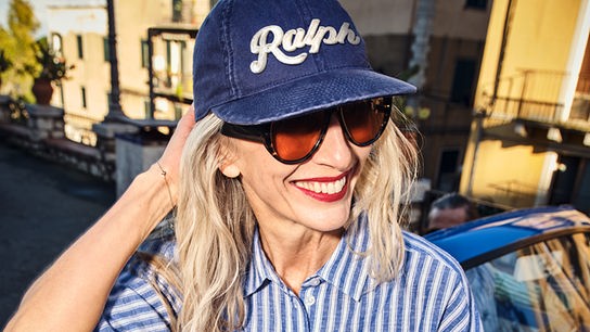 © Peek & Cloppenburg Eine blonde Frau mit Schirmkappe steht lässig gekleidet auf einer Straße vor einem blauen Auto.