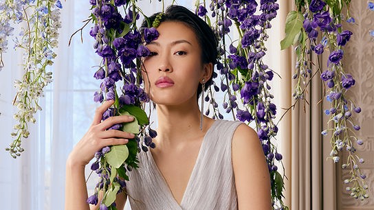 © Peek & Cloppenburg Eine junge Frau in einem sommerlichen Kleid steht in einem Innenraum, umrahmt von herabhängenden violetten Blüten.