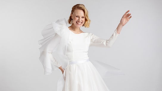 Eine Frau trägt ein voluminöses, weißes Brautkleid und posiert vor neutralem Hintergrund.