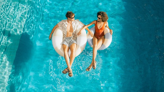 Ein Mann und eine Frau treiben entspannt auf hellen Schwimmreifen in einem türkisblauen Pool.