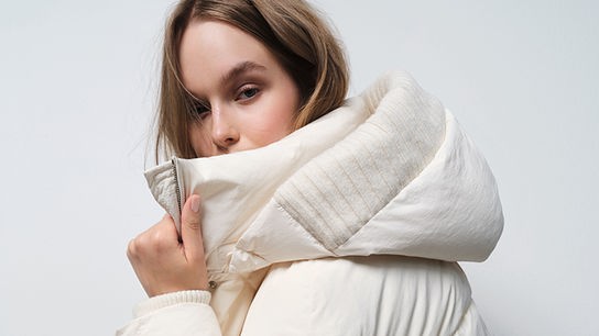 Eine Frau trägt eine helle, gepolsterte Winterjacke in der Farbe Pantone Cloud Dancer in einem schlichten Studio-Setting.