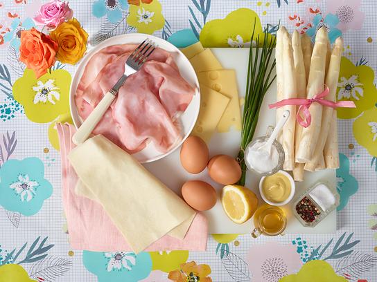 Verschiedene Zutaten wie Spargel, Schinken, Käse, Eier und Gewürze sind auf einem farbenfrohen Untergrund arrangiert.