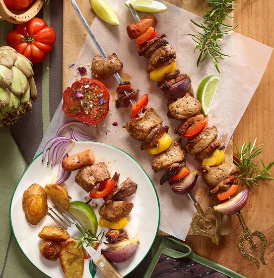 Auf einem Tisch liegen gegrillte Fleisch‑ und Gemüsespieße mit Kräutern, Kartoffeln und frischen Zutaten dekorativ angerichtet.