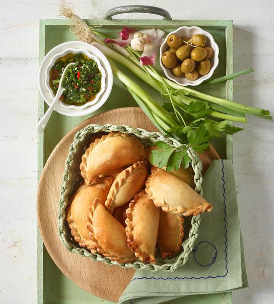 In einem Korb liegen goldbraun gebackene Empanadas, dazu Kräutersauce, Oliven und frisches Gemüse.