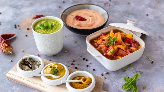 Mehrere bunte Dips und Schälchen mit Frucht- und Gemüsegerichten stehen dekorativ angerichtet auf einer hellgrauen Platte.