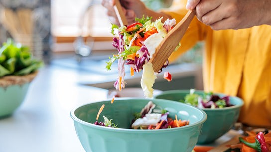 Eine Person mischt mit Holzlöffeln einen bunten Salat in einer Schüssel in einer hellen Küche.