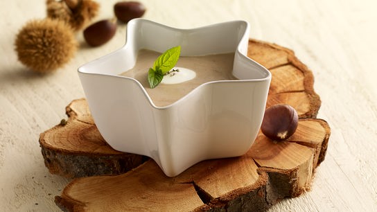 © APA-Images Cremige Maronisuppe in einem sternförmigen weißen Teller, dekoriert mit Minzblatt, auf einem Holzbrett serviert.