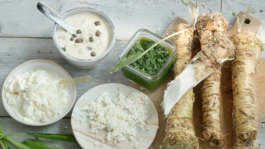 Frischer Kren mit Beilagen wie Topfen, Sauce und Kräutern liegt auf einem rustikalen Holztisch.