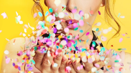© 123RF/artmim Eine Frau pustet buntes Konfetti aus den Händen vor gelbem Hintergrund.