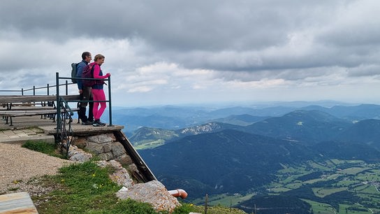 Christine Reiler und ein Mann stehen in Wanderbekleidung auf einer Aussichtsplattform und blicken über eine weite Berg‑ und Tallandschaft.