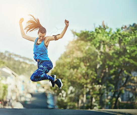 Eine sportlich gekleidete Frau springt energiegeladen in die Luft, vor einem sonnigen Straßen- und Naturhintergrund.