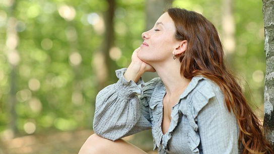 Eine Frau sitzt entspannt mit geschlossenen Augen an einen Baum gelehnt in einer grünen Waldumgebung.