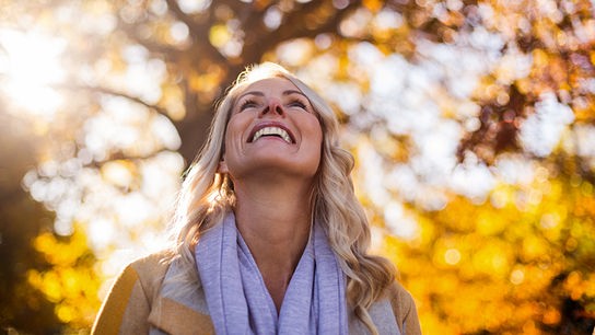 Eine lachende Frau steht im warmen Licht vor herbstlich gefärbten Bäumen.