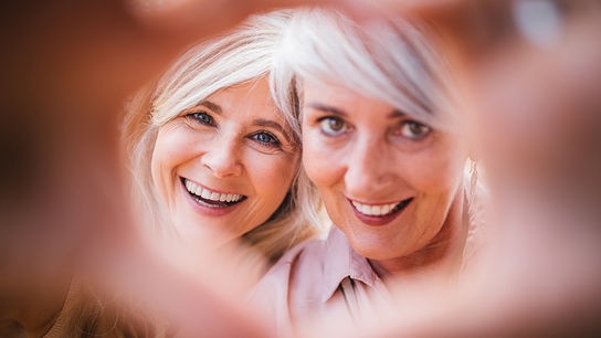 Zwei grauhaarige Frauen lächeln gemeinsam in die Kamera, die von Händen eingerahmt wird.