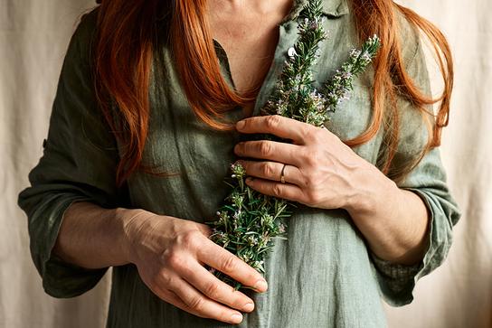 Eine rothaarige Frau hält mehrere blühende Rosmarinzweige mit den Händen an ihre Brust.