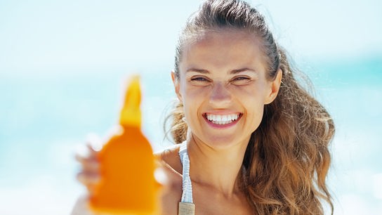Eine Frau im Bikini am Strand hält eine Sonnencreme in die Kamera, das Meer ist unscharf im Hintergrund zu sehen.