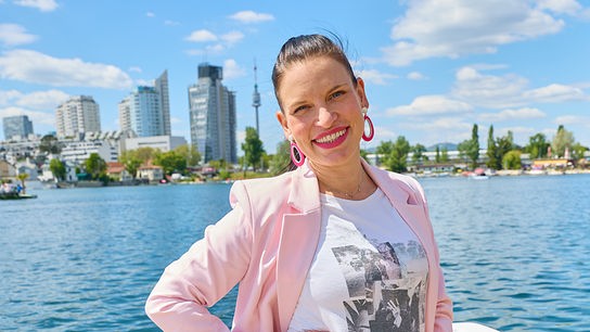 Conny Kreuter steht lächelnd auf einem Boot an der Donau, im Hintergrund die Skyline von Wien.