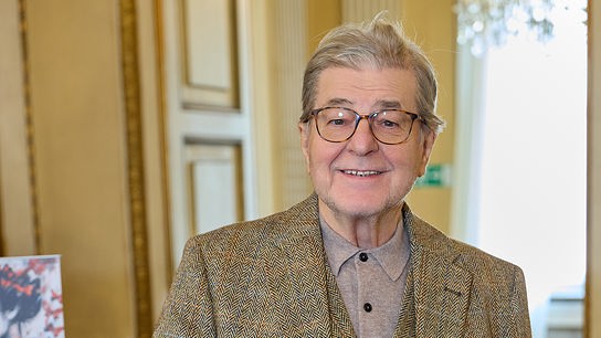 Ein lächelnder Erwin Steinhauer steht in einem eleganten Raum neben einem Flügel und trägt ein braunes Tweed-Sakko.