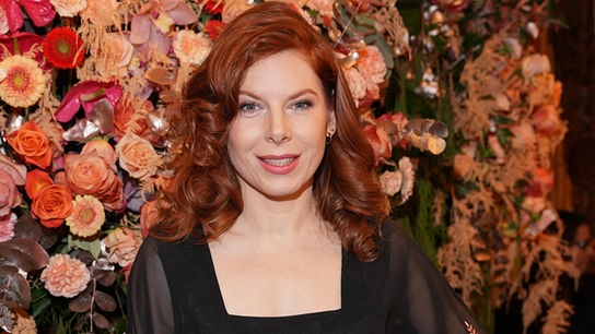 © ORF/Roman Zach-Kiesling Teresa Vogl posiert in einem schwarzen Kleid vor einer üppigen Blumenwand.