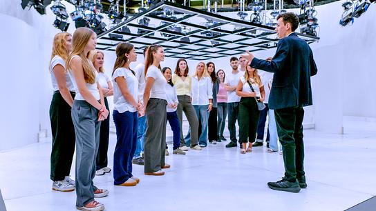 Eine Gruppe junger Erwachsener steht in einem hellen Studio in zwei Reihen und hört einem Mann zu.