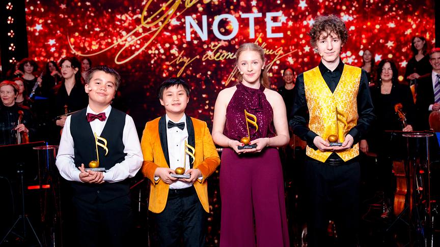 Die vier Gewinner:innen der 10. Goldene Note Lukas Maierhofer, Jayden King, Linda Gittmaier und Adam Benold stehen auf einer festlich beleuchteten Bühne und halten goldene Noten-Trophäen vor dem Schriftzug „Goldene Note“, während im Hintergrund ein Orchester zu sehen ist. 