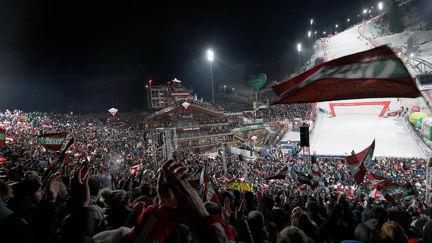 © GEPA_Hans Oberländer Ein dicht gedrängtes Publikum schwenkt Fahnen und jubelt bei einem nächtlichen Ski-Weltcup-Rennen in Schladming im hell erleuchteten Zielstadion.