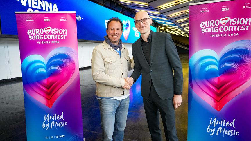 ESC-Executive-Producer Michael Krön und Thomas Waldner (Geschäftsführer der Wiener Stadthalle) geben sich die Hand vor Roll‑up‑Bannern des Eurovision Song Contest 2026 in der modernen, beleuchteten Wiener Stadthalle.
