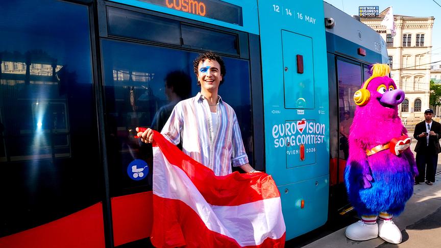 COSMÓ steht vor der „TANZSCHEIN“-Sonderbim mit Eurovision‑Beschriftung und hält eine rot‑weiß‑rote Flagge, im Hintergrund steht das bunte Maskottchen Auri.