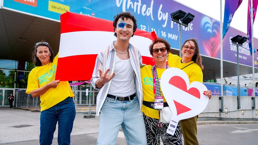 COSMÓ und drei Volunteers posieren vor der Wiener Stadthalle mit österreichischer Flagge und Herzsymbolen in Fan‑Outfits.