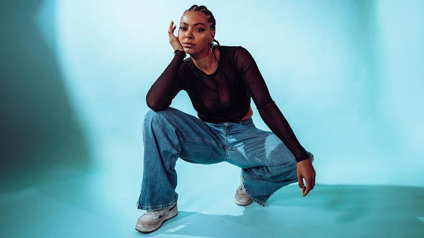 © ORF/FM4/suna.films NENDA hockt vor hellem, türkisfarbenem Hintergrund in weiten Jeans, weißen Sneakers und einem schwarzen, durchsichtigen Oberteil, mit einer Hand seitlich am Kopf und der anderen nach unten gestreckt.