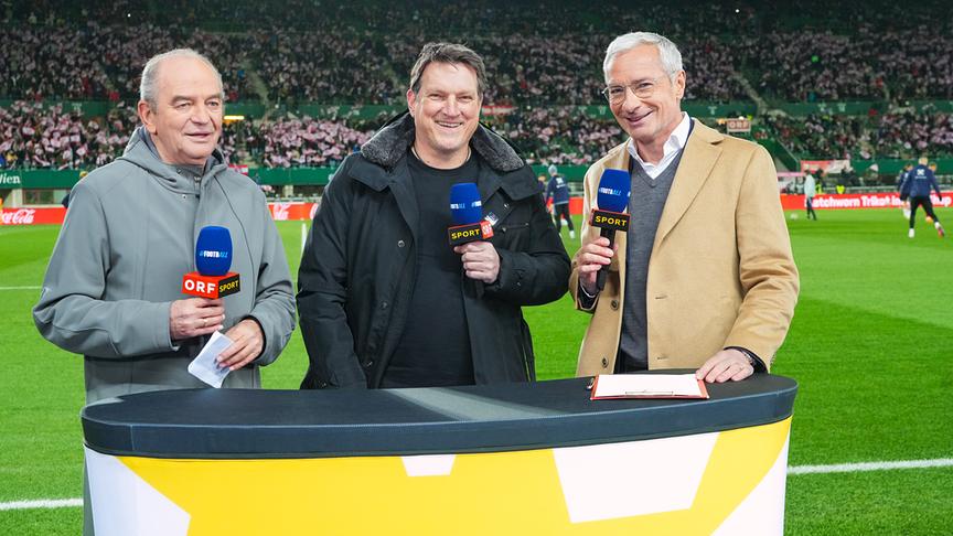Herbert Prohaska, Andreas Herzog und Rainer Pariasek stehen mit ORF-Sport-Mikros hinter dem Moderatorenpult vor dem Fußballfeld in einem voll besetzten Fußballstadion.