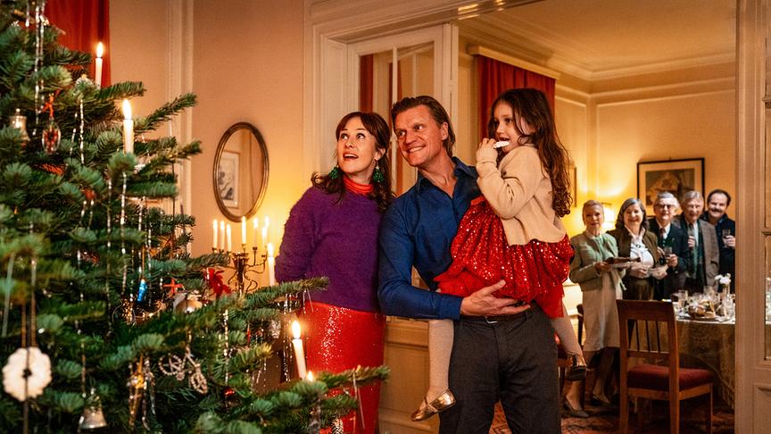 Steffi, Andi und Alma stehen vor einem hell erleuchteten und geschmückten Christbaum in einer Altbauwohnung.