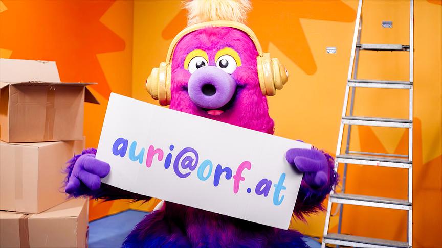AURI, ein pinkfarbenes, kopfhörertragendes Fantasiewesen, hält vor Umzugskartons und einer Leiter ein Schild mit der Aufschrift „auria@orf.at“.
