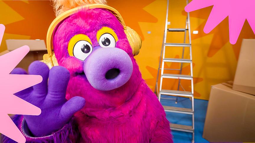 AURI, ein pink-violettes Maskottchen mit goldenen Kopfhörern, posiert in einem orangefarbenen Studio vor einer Leiter.