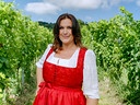 Barbara Karlich in rotem Dirndl steht zwischen grünen Weinreben unter blauem Himmel.
