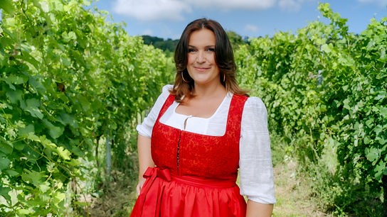 Barbara Karlich in rotem Dirndl steht zwischen grünen Weinreben unter blauem Himmel.