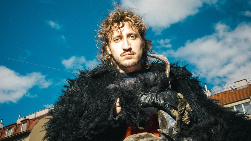 © ORF/www.berniwagner.at/Christopher Glanzl Berni Wagner im schwarzem Fellmantel und einer Monstermaske in der Hand steht vor einer Häuserzeile unter einem Wolkenhimmel.