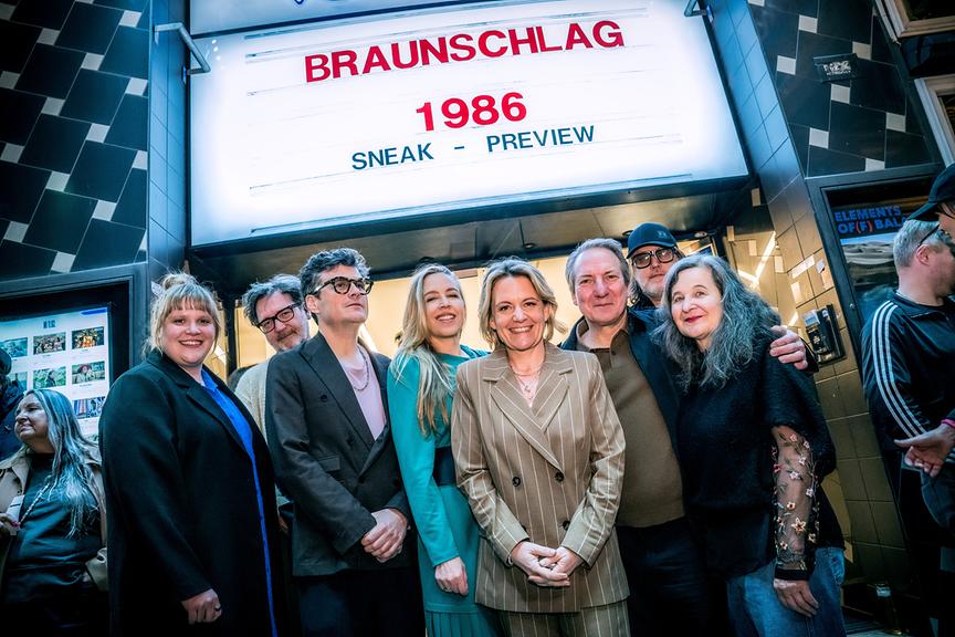 © ORF/Thomas Jantzen Stefanie Reinsperger, Regisseur und Produzent David Schalko, Manuel Rubey, Nina Proll, ORF-Programmdirektorin Stefanie Groiss-Horowitz, Robert Palfrader, Nicholas Ofcazrek und Maria Hofstätter posieren abends vor einem Kinoeingang, über dem auf einer Leuchttafel „Braunschlag 1986 – Sneak Preview“ zu lesen ist.