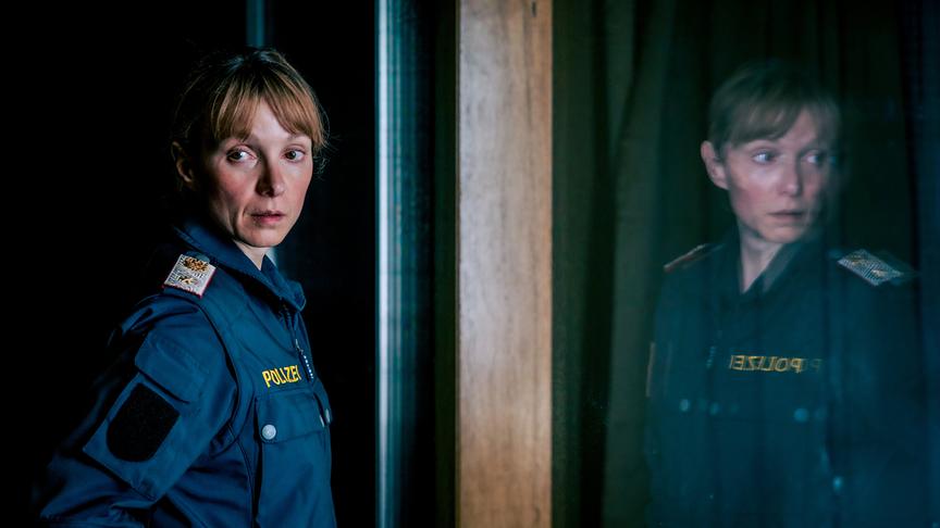 © ORF/Mona Film/Viktoria Herbig Julia Koschitz in Polizeiuniform steht vor einer dunklen Glasfläche, in der sich ihr Spiegelbild zeigt.