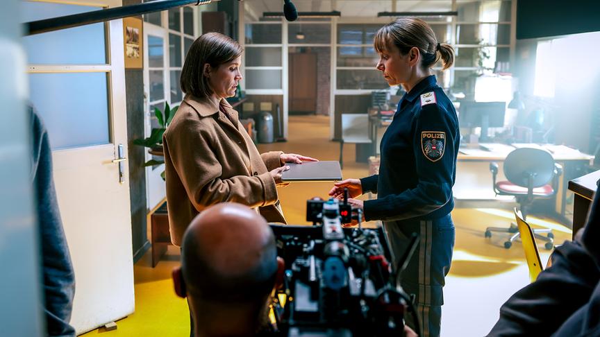 © ORF/Mona Film/Viktoria Herbig Julia Koschitz in Polizeiuniform steht in einem Büro und nimmt von Mona Kospach eine Mappe entgegen, während eine Filmkamera und ein Mikrofon sichtbar sind.