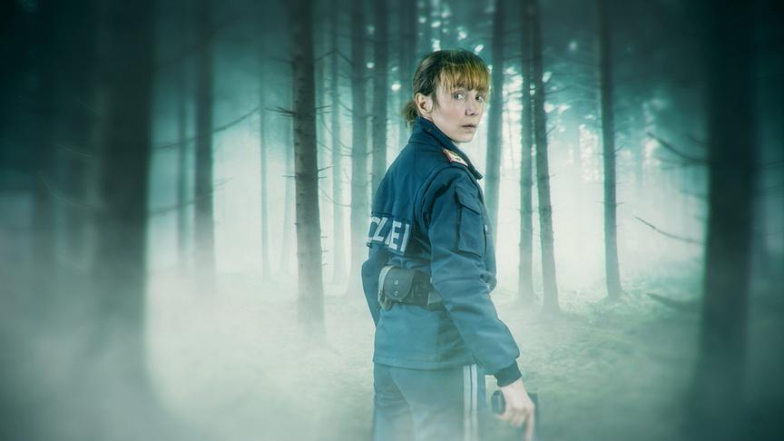 Julia Koschitz als Polizistin Pia Kraus steht mit einer Pistole in einem nebligen Wald und blickt angespannt über die Schulter.