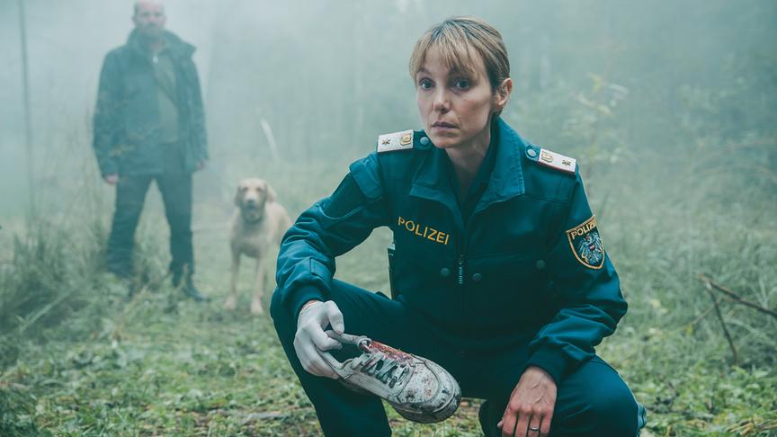 © ORF/ARD Degeto/Mona Film/Victoria Herbig Im Moor: Polizistin Pia Krauss hält einen blutverschmierten Turnschuh in der Hand, der möglicherweise der Vermissten gehört. Hinter ihr steht Gruber mit seinem Hund, der den Schuh gefunden hat.