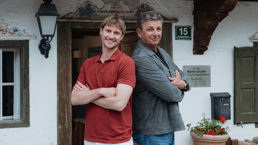 Frédéric Brossier und Hans Sigl stehen mit verschränkten Armen Rücken an Rücken vor dem Eingang eines Bauernhauses bei den „Der Bergdoktor“-Dreharbeiten zur Jubiläumsstaffel