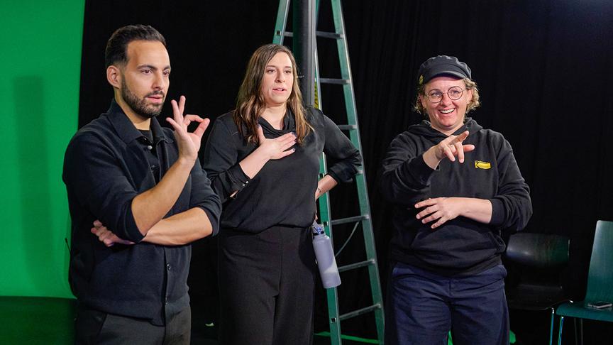 Amanda Jovanovic mit dem Coaching Team Delil Yilmaz und Magdalena Schramek stehen in einem Studio und führen mit ihren Händen Gesten aus.